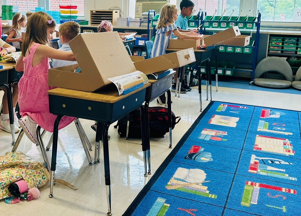 elementary students opening school supply boxes on desks in front of a colorful rug