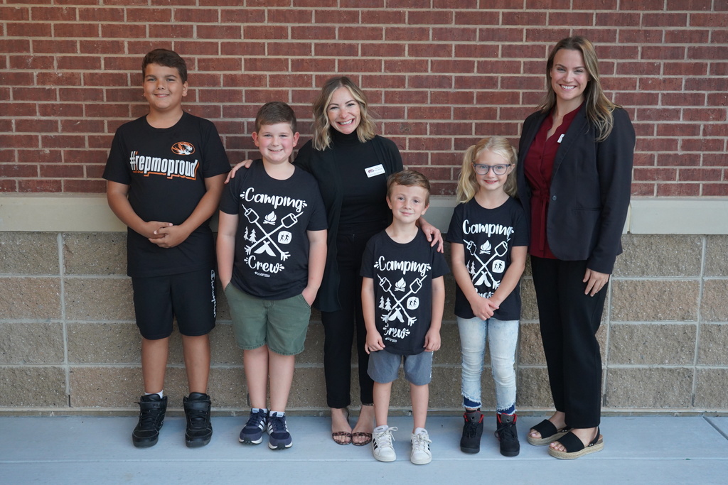 Students in black t-shirts stand and smile against a red brick wall with their principals