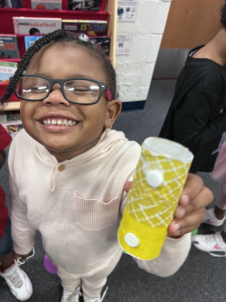 A kindergarten student grins while showing her "bus" that is yellow & made from cardboard and buttons.