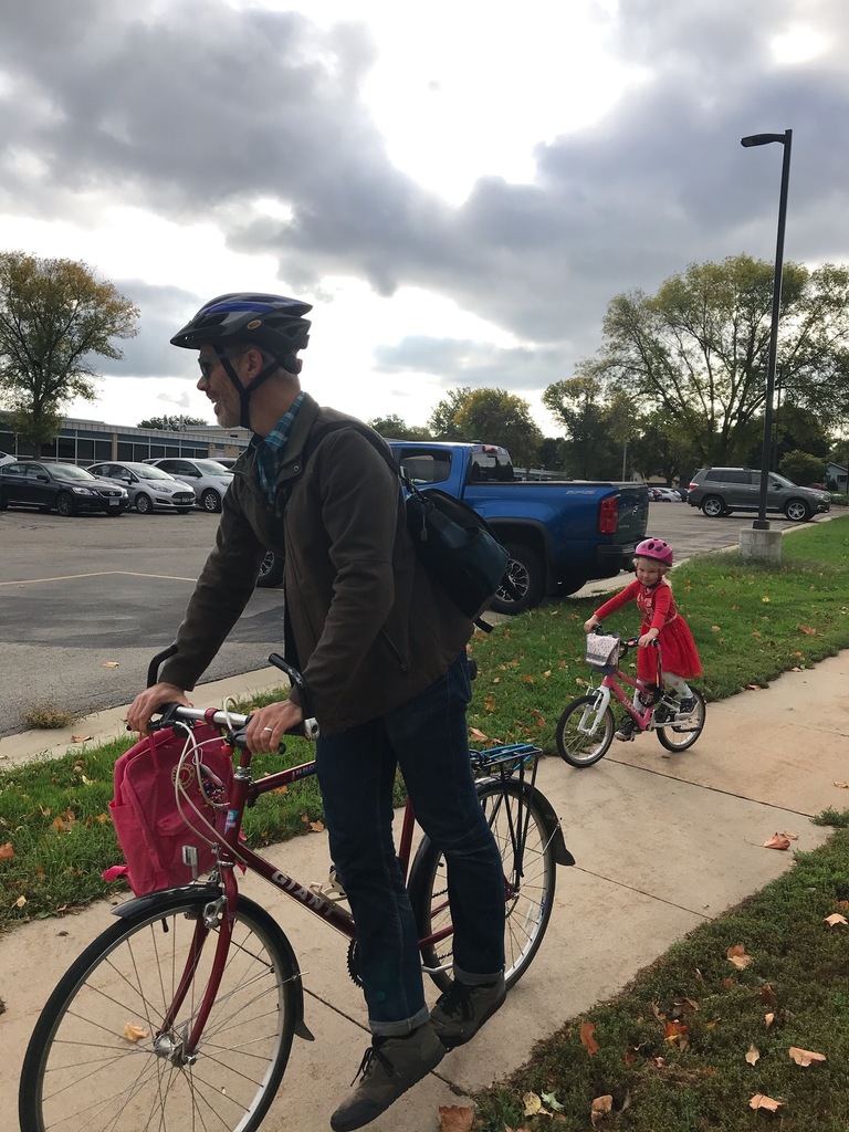 Dad walking his bike on the sidewalk with daughter riding bike.