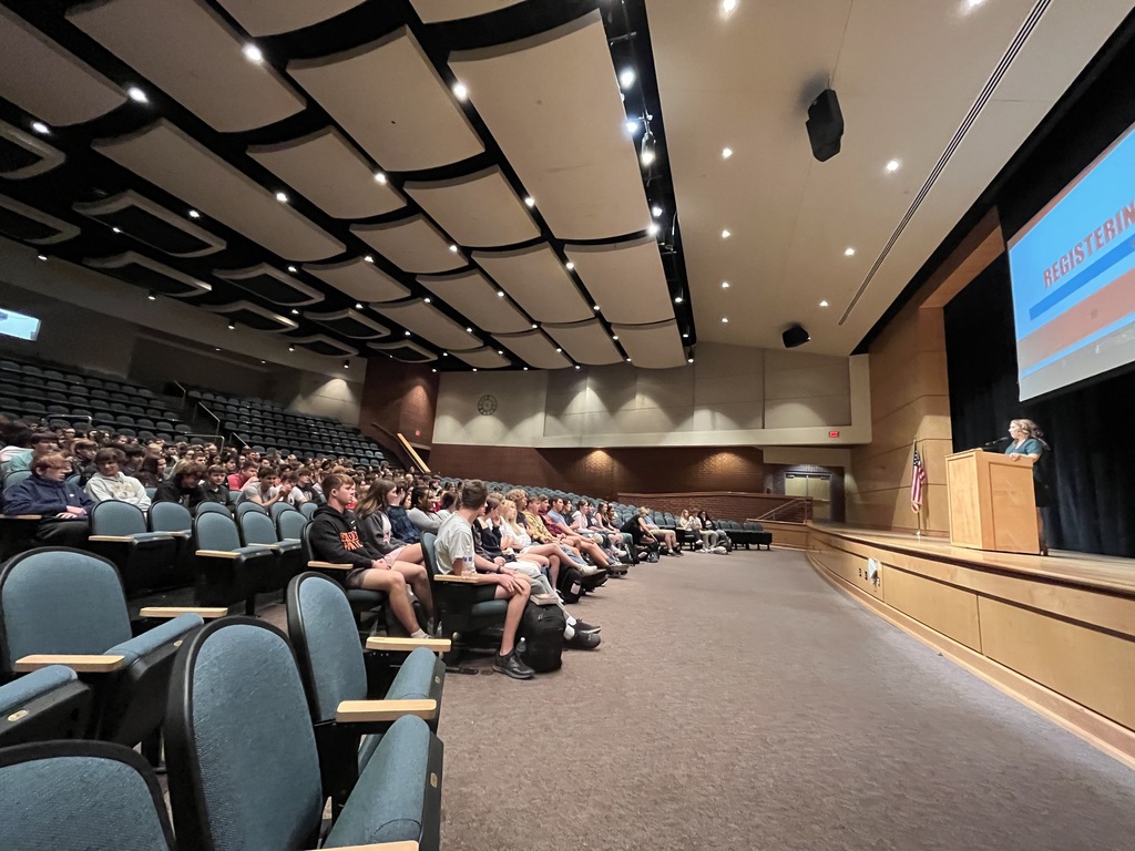 A woman speaks to a large group of high school students in an auditorium.