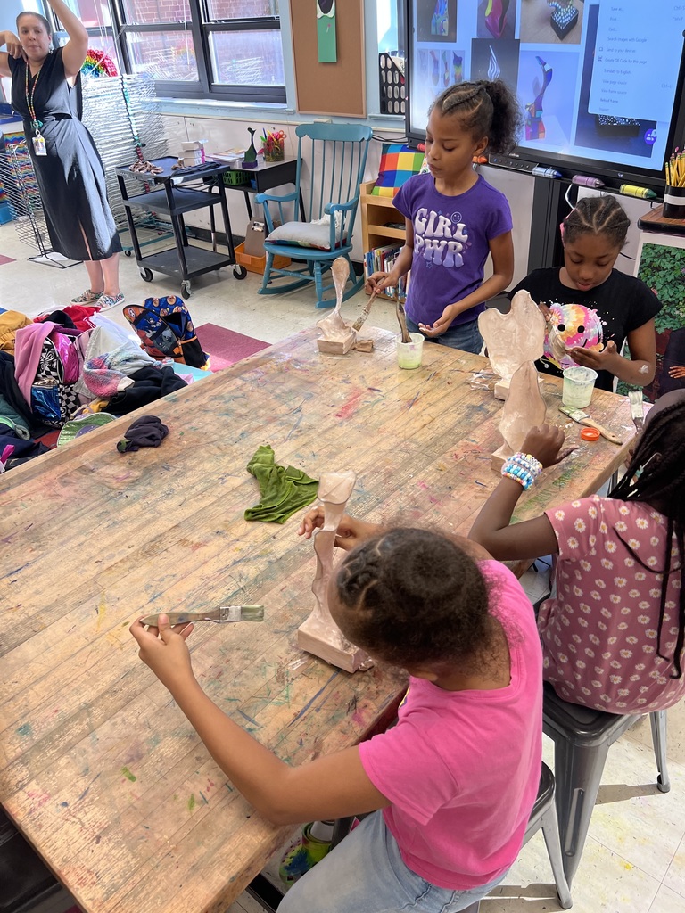 Four students paint their sculptures at a wooden table.