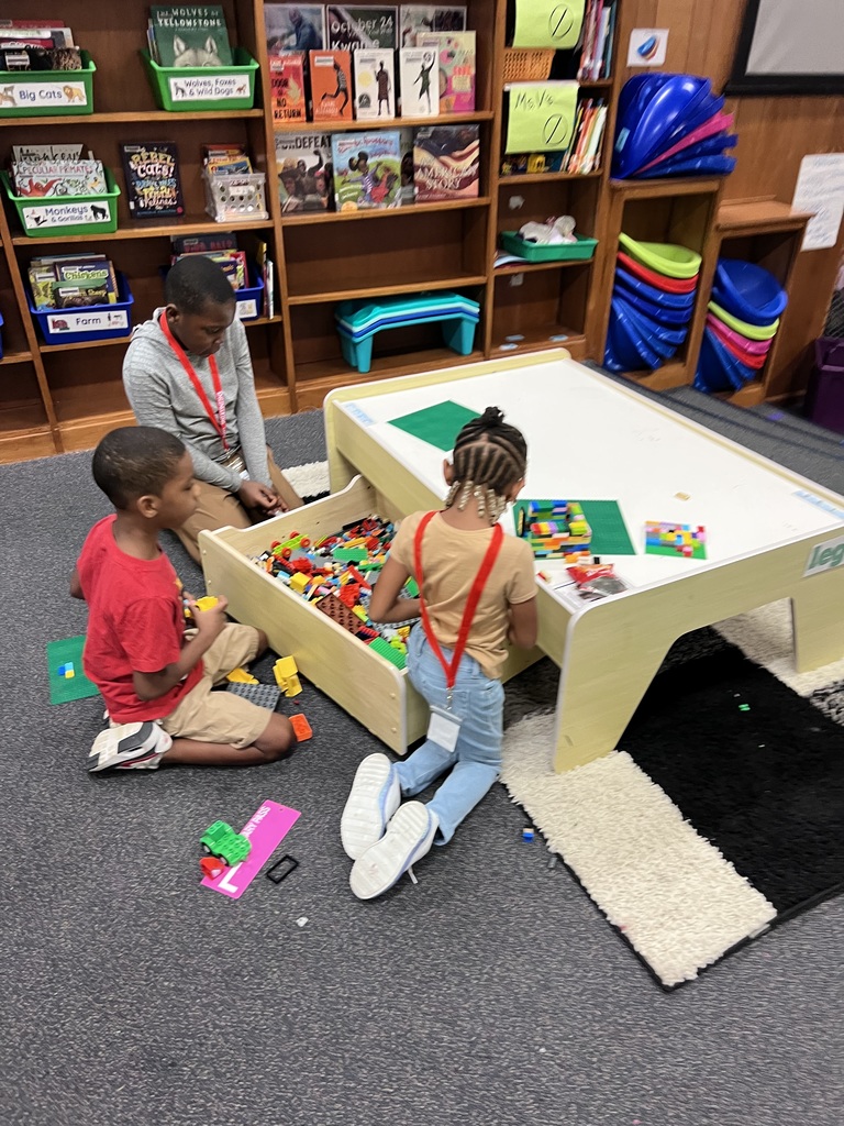 Three students across grade levels share the Lego table.