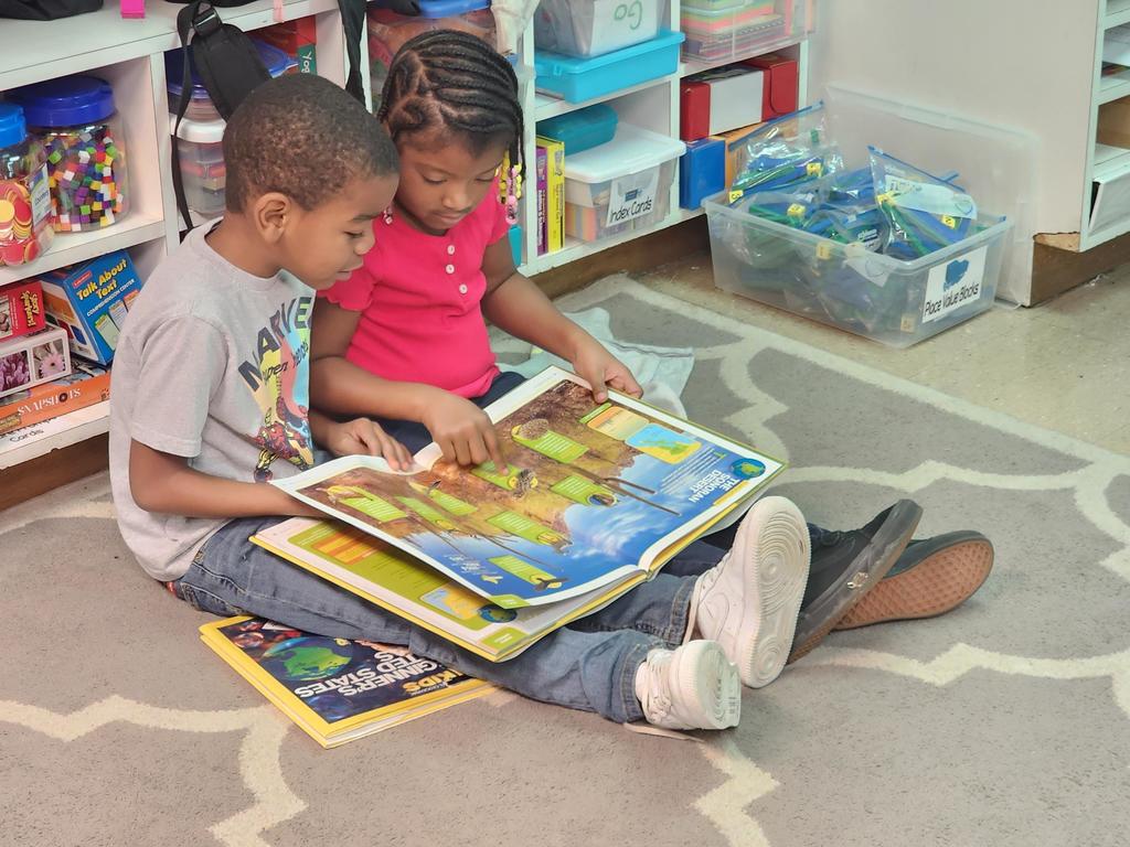 Two students browse pages in a large illustrated atlas.
