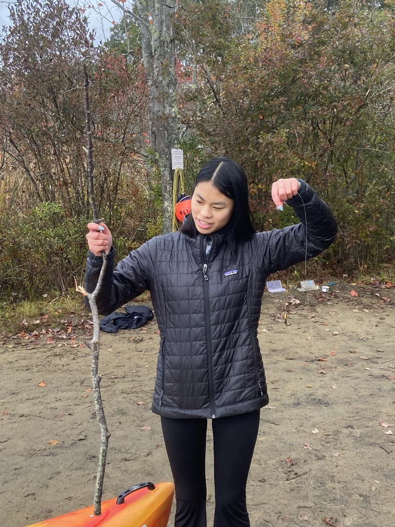 CE Student in a black jacket and black leggings in the woods holds a stick "fishing pole" near an orange kayak