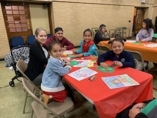 Four students and there parents sitting around rectangular tables with red table cloths.  The sutdents are coloring.