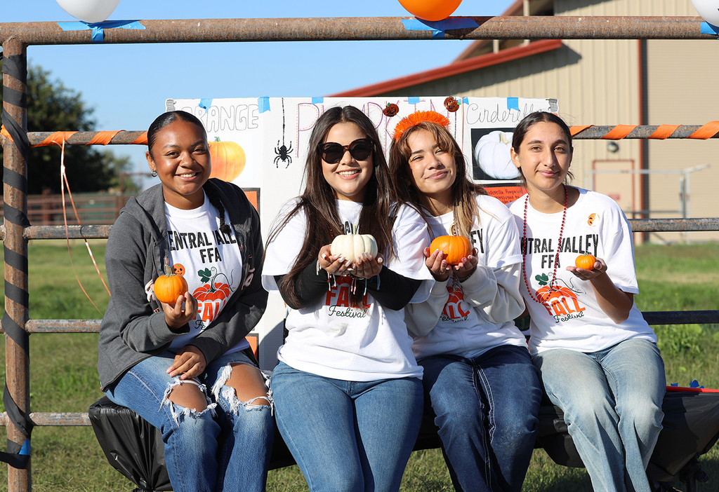 Four female high school students sitting on fence rails holding pumpkins