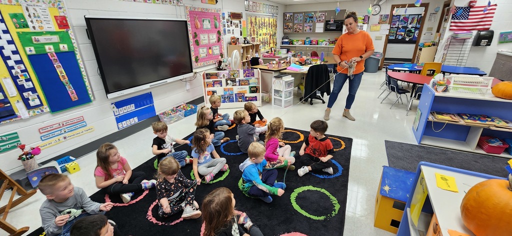 An adult is standing in a 4K classroom, holding a string that a student sitting on the carpet is holding. Other students sitting on the carpet also have string in their hands.