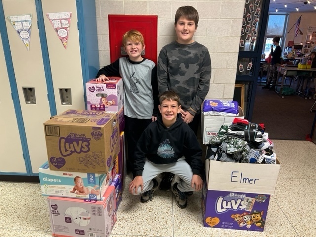 3 male elementary students wearing black and grey are standing between boxes of diapers and socks.