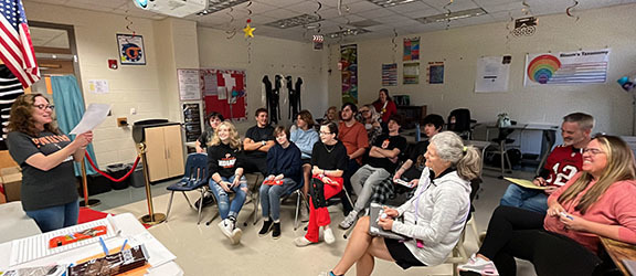 A group of students and adults listen to a woman announce the awards of a film festival.