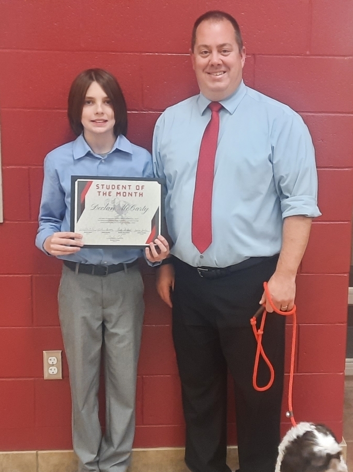 one student and one teacher in blue shirts stand in front of a red wall. Student is holding a certificate.