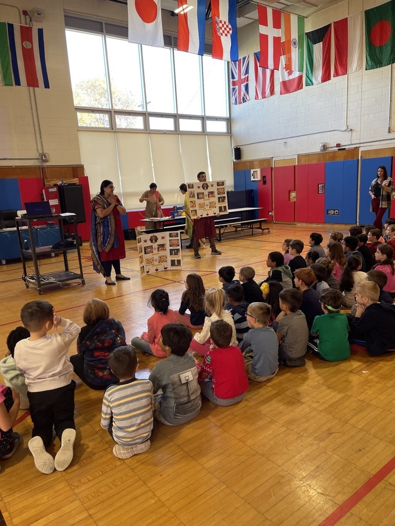 several classes of students sitting in the school gymnasium watching parents give a presentation on Diwali