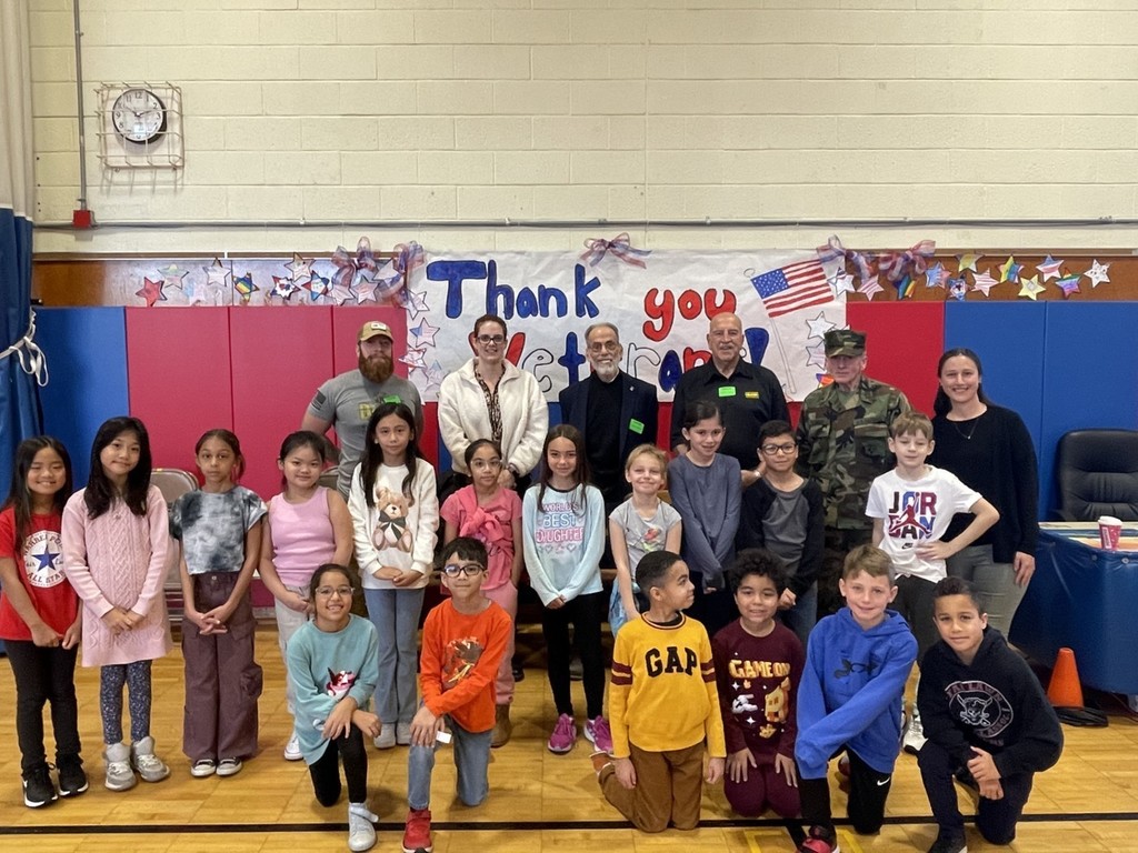 3rd grade class standing in school gymnasium with their teacher and 5 veterans