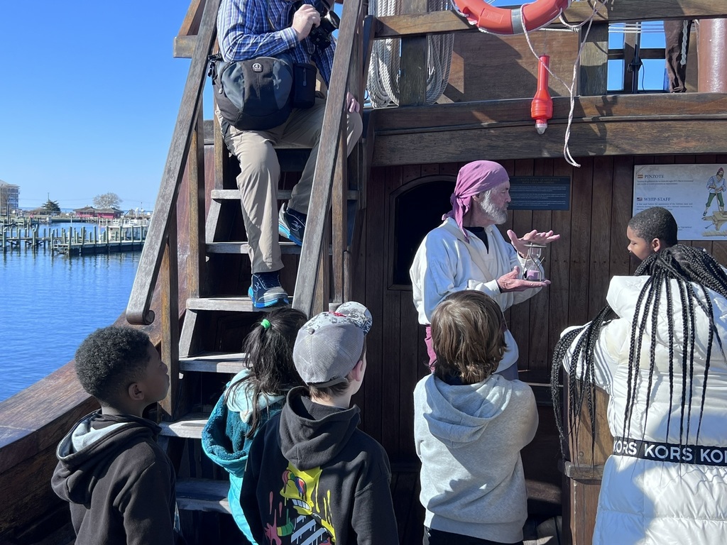 ship mate holding a hour glass and explaining to a student how they kept track of work and rest times