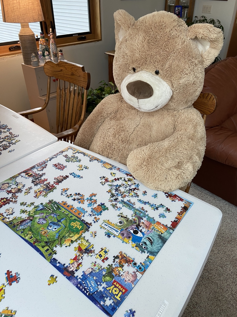 Teddy Bear sitting at table looking at puzzle.