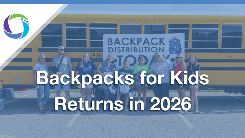 A promotional image featuring eight people—adults and children—standing in a parking lot in front of a school bus. They are positioned behind a white banner that says "Backpack Distribution Today" with a backpack graphic. A blue-tinted overlay covers the image with large white text centered that says "Backpacks for Kids Returns in 2026." The top left corner features a circular logo with purple, green, and blue segments.