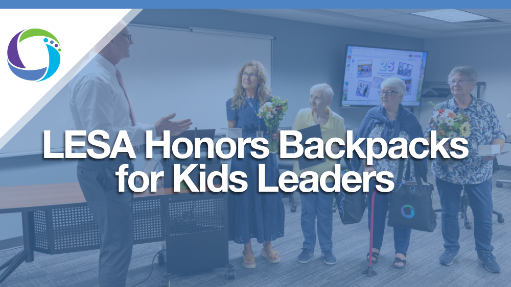 Graphic featuring the Livingston Educational Service Agency (LESA) logo in the upper left corner over a blue-tinted photo. In the image, a man in a dress shirt speaks to four women, some holding flowers, gathered in a presentation room. Bold white overlay text reads "LESA Honors Backpacks for Kids Leaders."