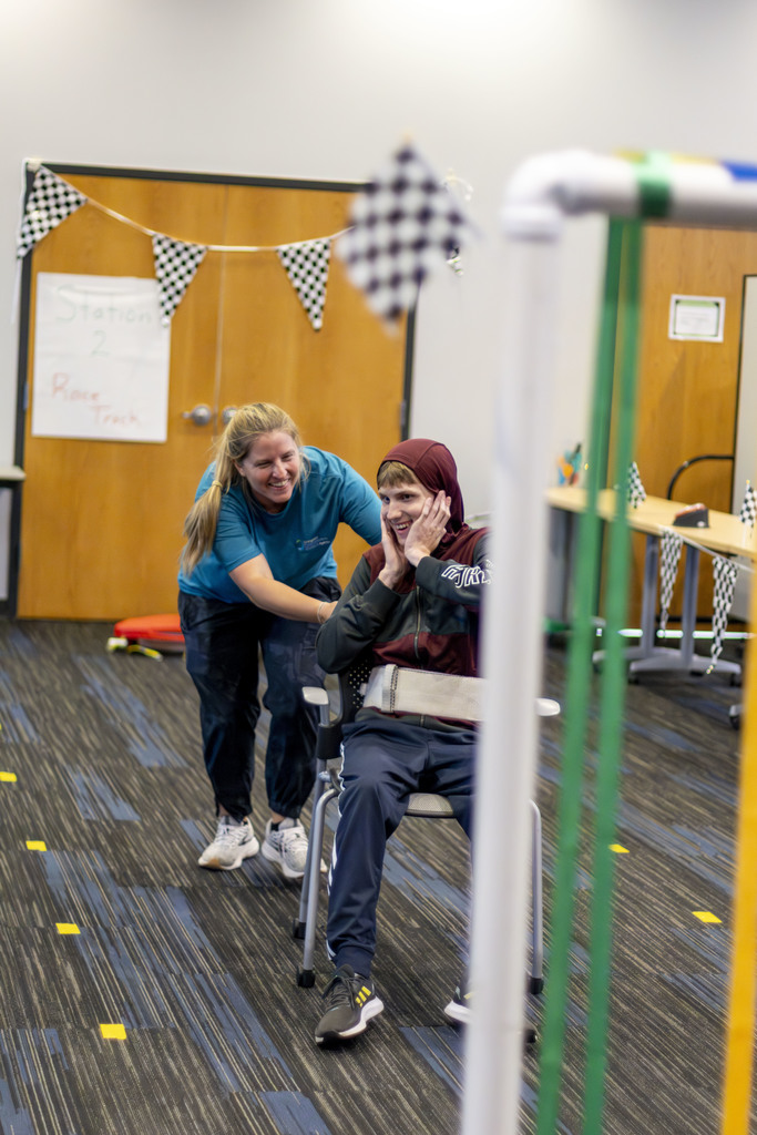 Smiling boy being pushed in chair by smiling teacher. 