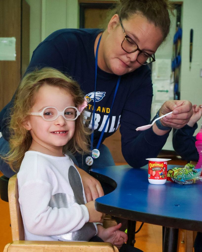Child posing for photos while the teacher tries to feed her.