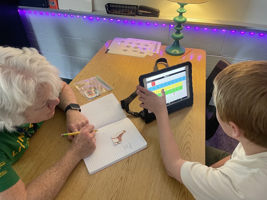 Student and teaching assistant sit at table. Student is using a keyboard, while the assistant scribes.