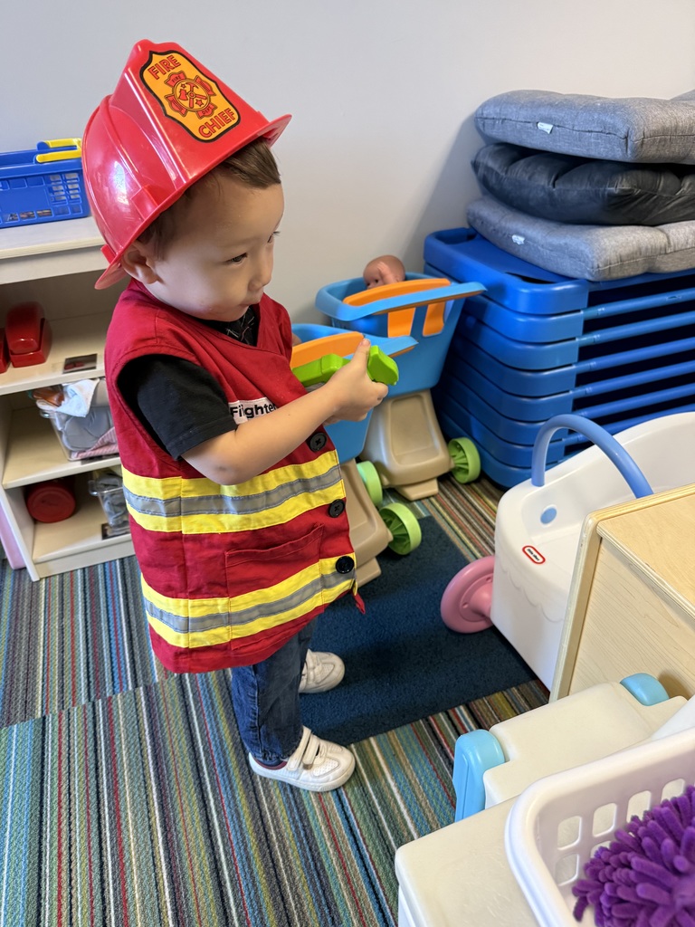 A young child dressed as a firefighter holds toy tools while playing in a classroom area.