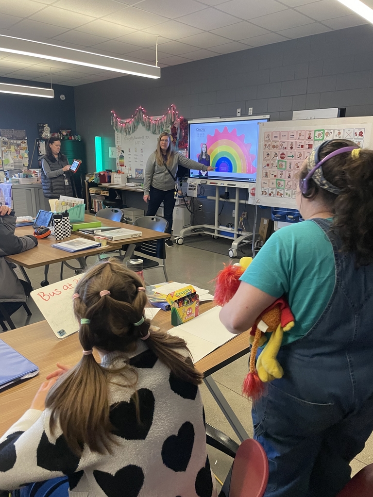 An instructor points to a screen showing a rainbow and the word "Circles" while students watch in a modern classroom.