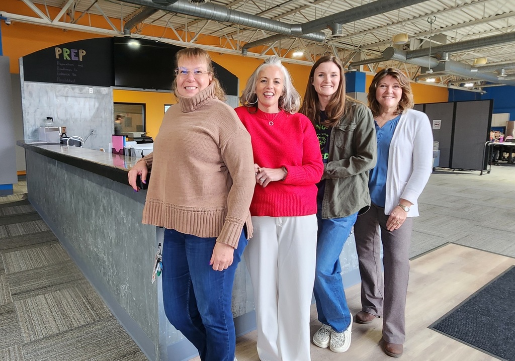 Four educators standing and smiling together at the front desk in a business-like school setting.