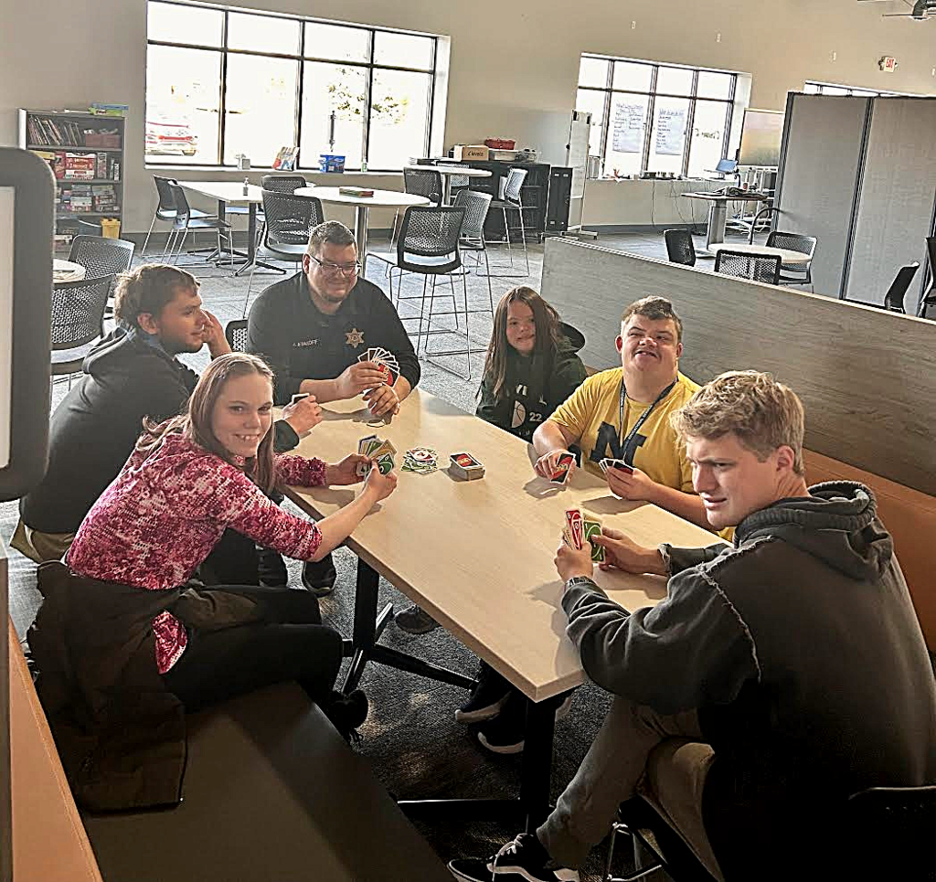Five young adults sitting at a booth, playing a game of Uno with a deputy sheriff.