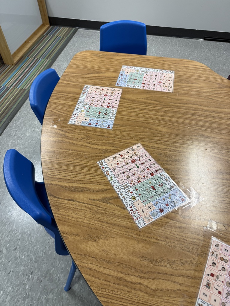 A classroom table with laminated picture communication boards taped at each seat for children to use during activities.