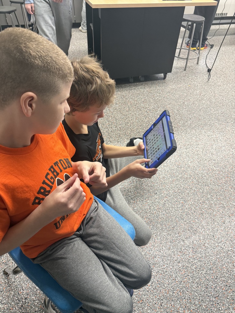 Two students sitting and kneeling on the floor. One student modeling words on a communication device. 