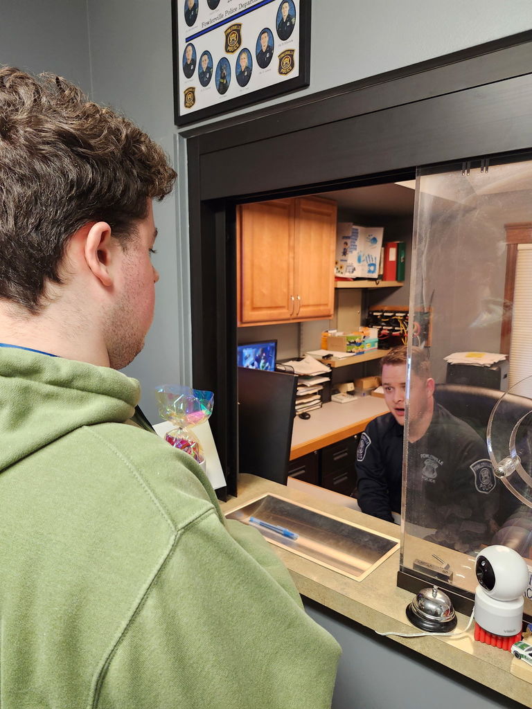 A young adult male standing on front of a counter with a male police officer sitting behind the counter.