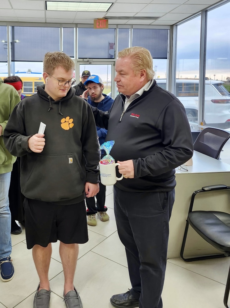 A young adult male standing with an adult male with a mug in his hand at a dealership.