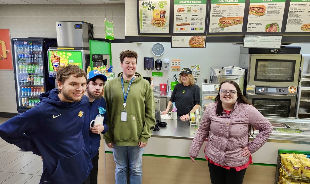 Three young adult males standing with a young adult female in front of a counter. A female worker is standing behind the counter of a restaurant.
