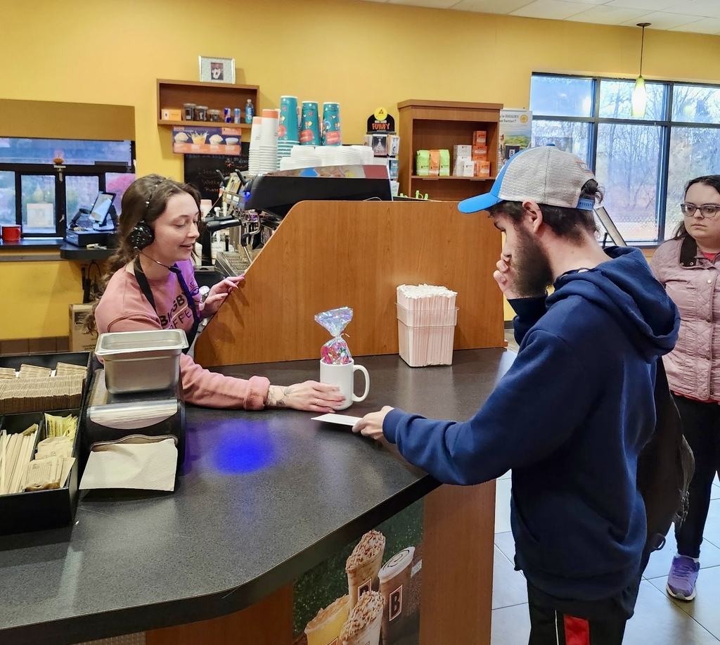 A young male adult handing a coffee mug filled with goodies and an envelope to a female adult in a coffee shop.