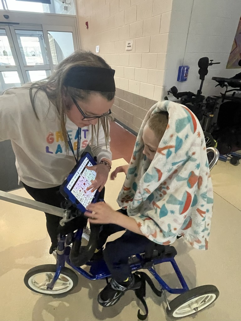 Student sitting on the bike with an ipad attached. Teacher is talking to the student. 
