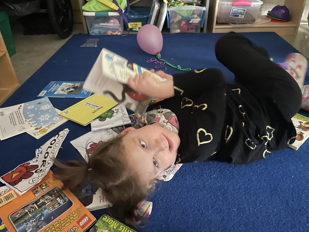 Student laying on the ground hold a book. There are additional books all around her.