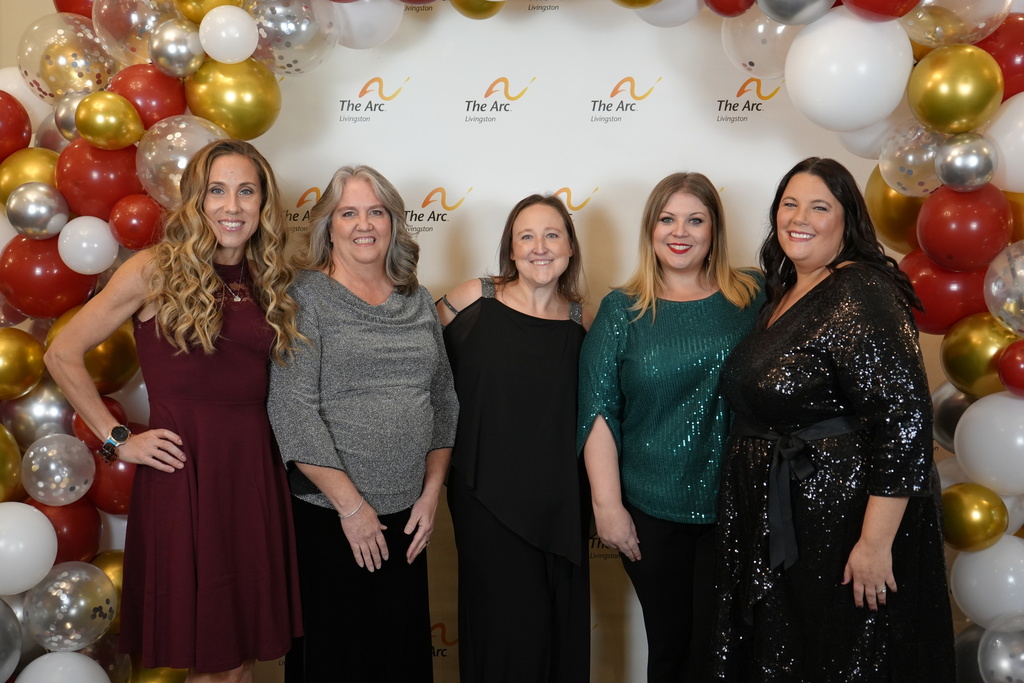 Five women posing together in front of a logo background with a balloon arch.