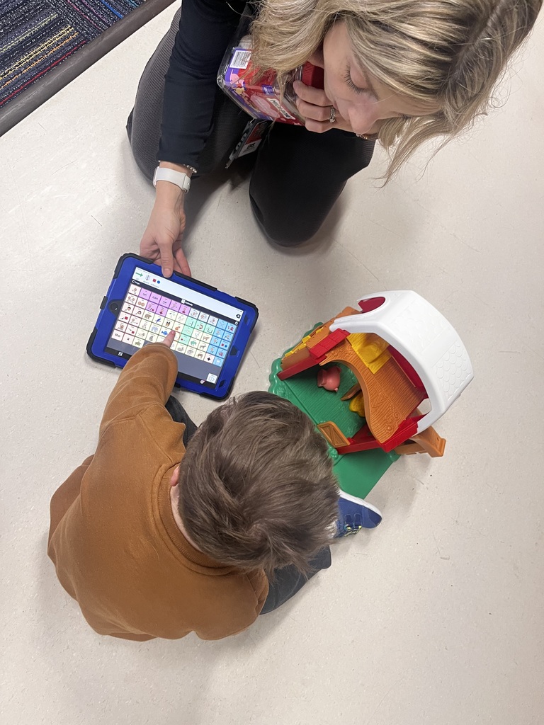 Photo from above of a female teacher holding a communication device and a student touching the device while playing with a toy barn.