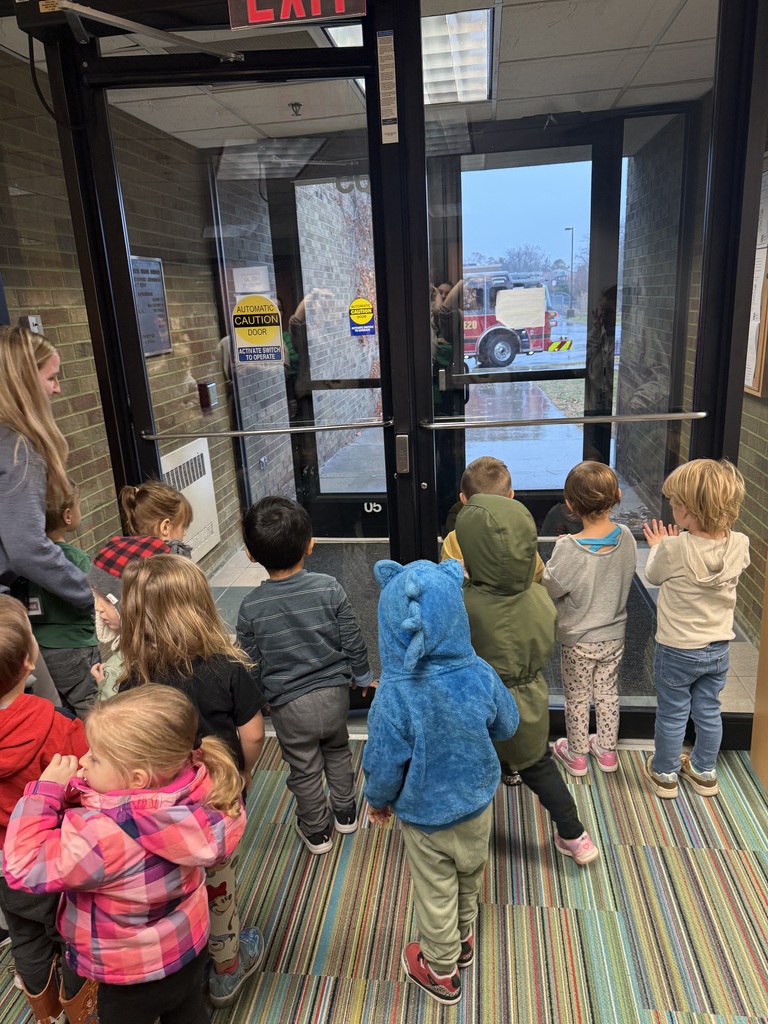 Children stand at a glass door watching a visiting fire truck outside.