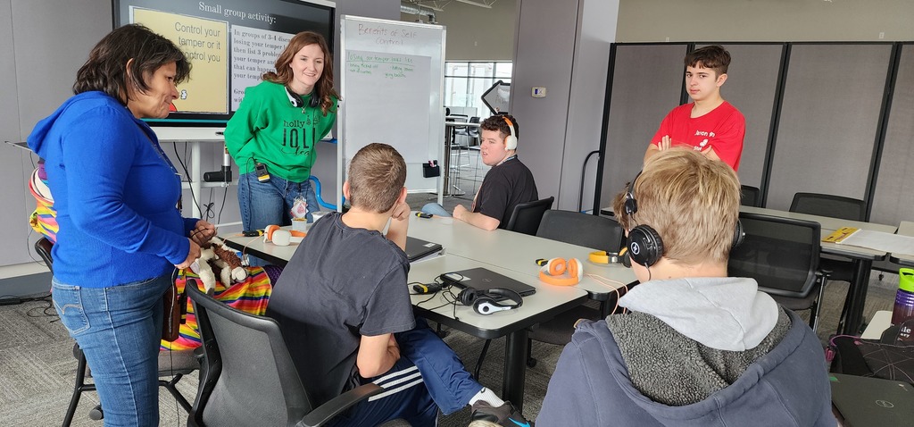 Two teachers standing, one adult male and three adult male learners sitting  around a table, having a discussion.