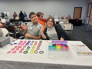 2 people sitting at a table selling handmaid bracelets 