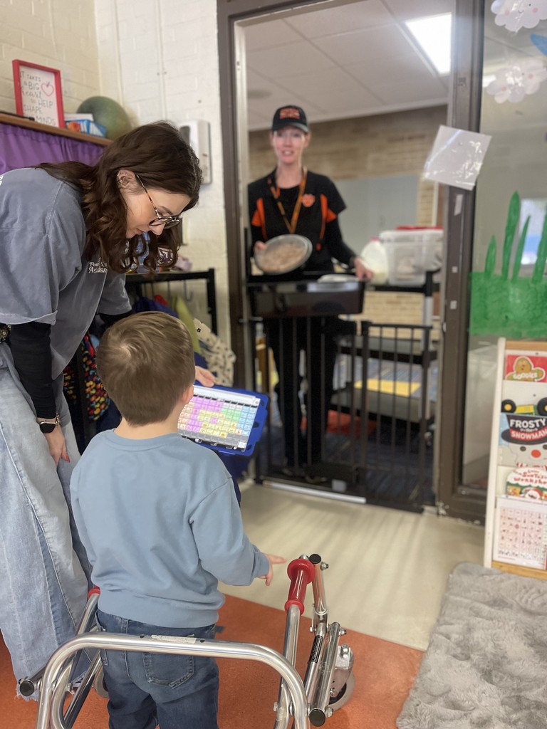 A young boy using a walker and a communication tablet interacts with a cafeteria worker in a doorway while a staff member assists him.