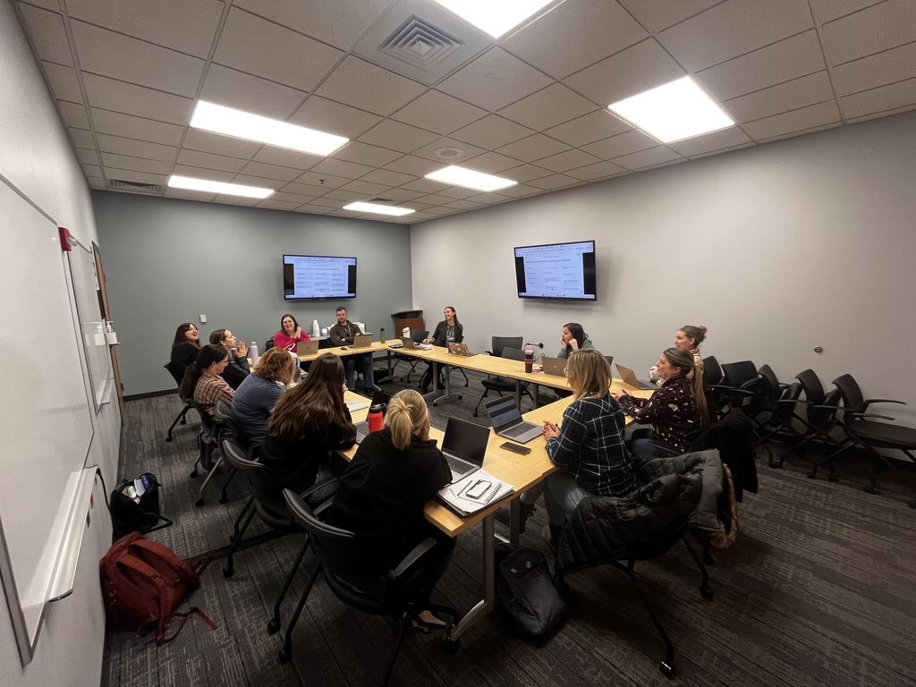 A group of colleagues sitting around U-shaped tables with laptops open during a collaborative meeting. 