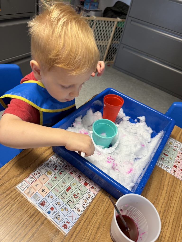 Child scooping colored snow in a sensory bin using cups and tools at a classroom table.