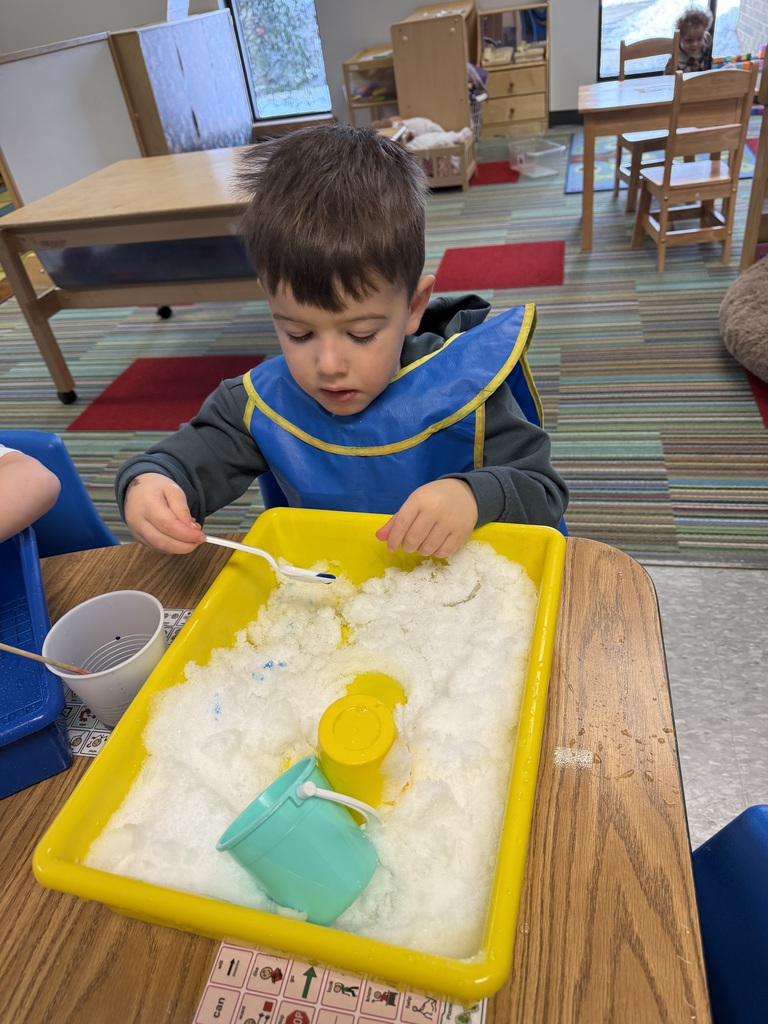 Child scooping colored snow in a sensory bin using cups and tools at a classroom table.