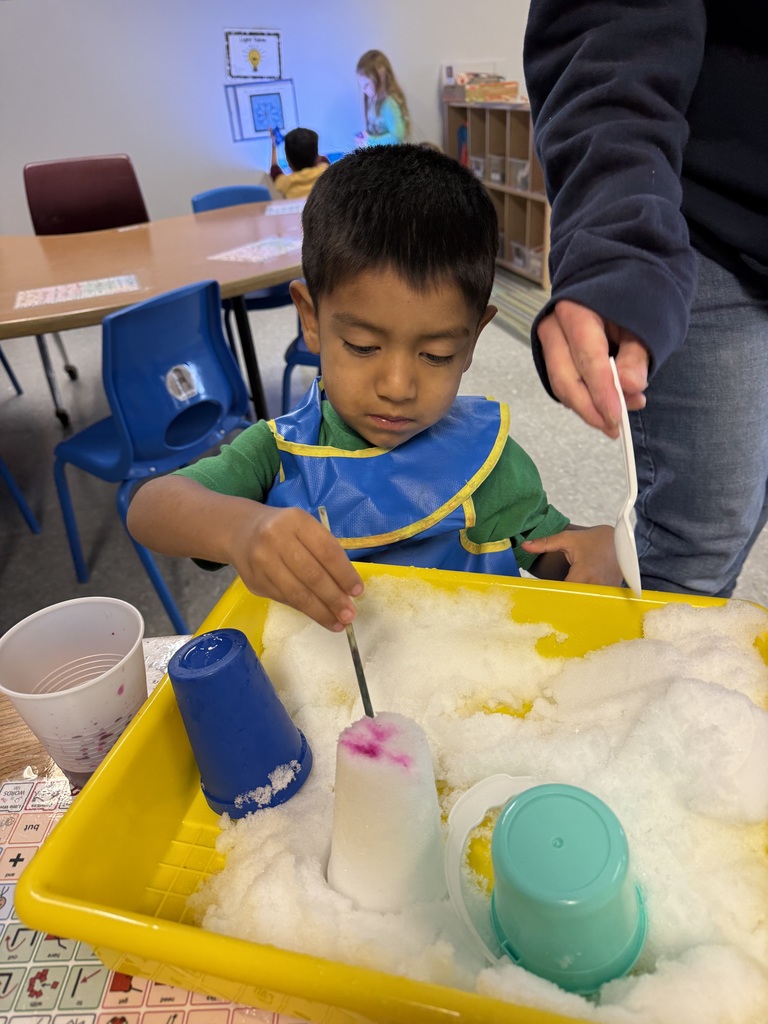 Child scooping colored snow in a sensory bin using cups and tools at a classroom table.