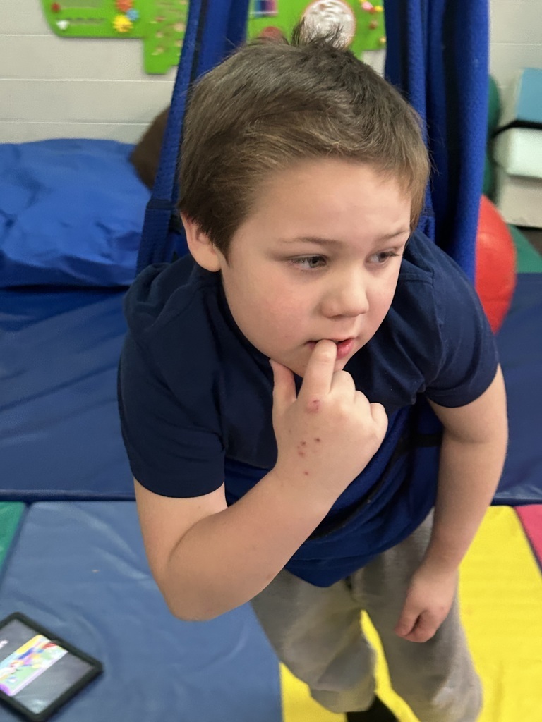 A young child hangs in a blue sensory swing, touching their lips thoughtfully, surrounded by colorful gym mats and equipment.
