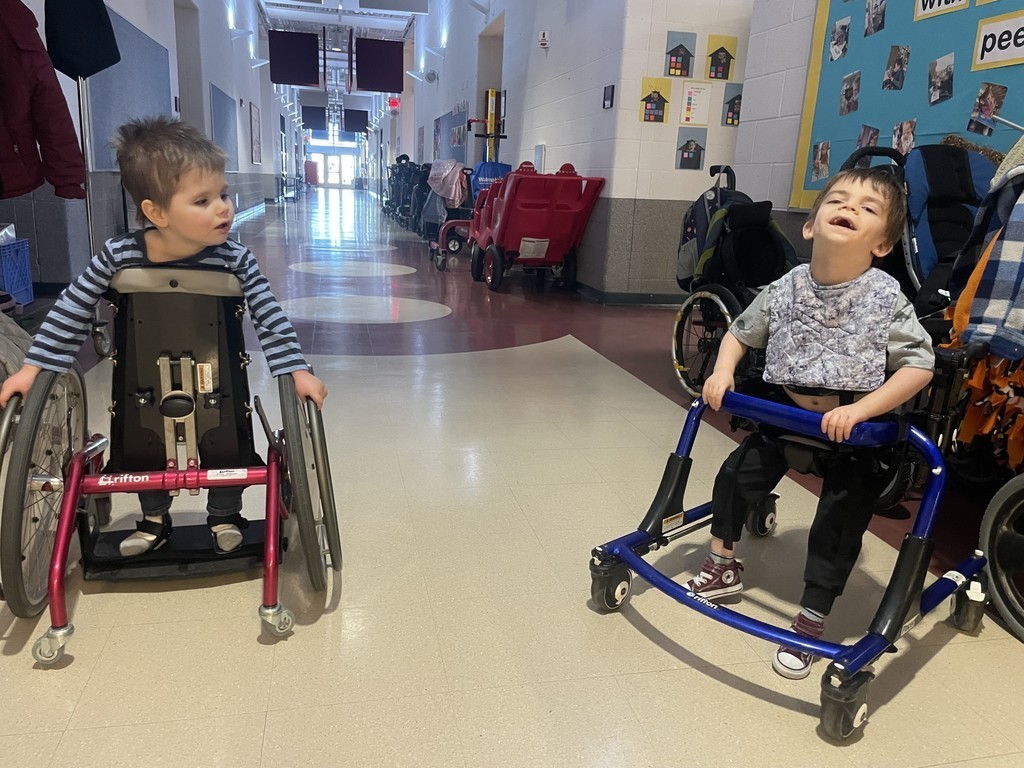 Two young learners in their adaptive devices in the hallway.