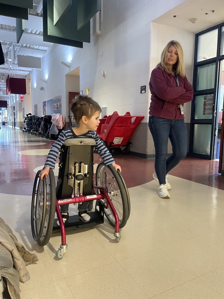 Student in mobile stander with teacher assistant in the hallway.