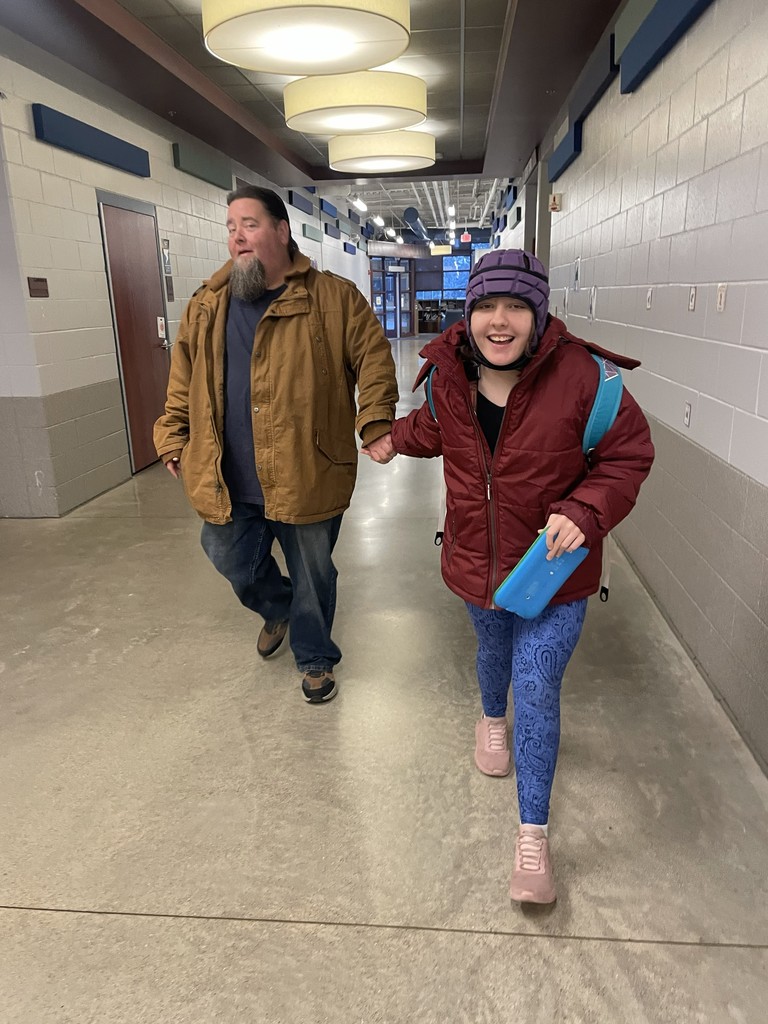 A student with a huge smile is holding hands with an assistant walking down the hall.